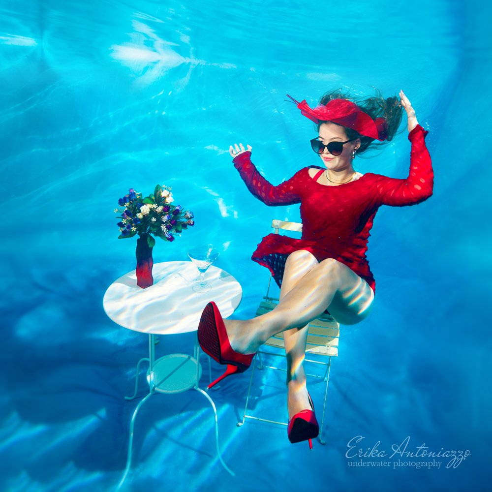 Onderwaterfoto model, jonge vrouw in rode jurk, hoed en schoenen, aan tafeltje met bloemen